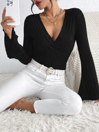 Woman sitting and taking a selfie while wearing a black long-sleeved ribbed knit top with a plunging V-neckline and flared sleeves, paired with white pants.