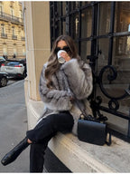 Woman sitting on a ledge outdoors wearing a grey faux fur coat, black top, black jeans, black boots, sunglasses, and gold earrings, holding a coffee cup.