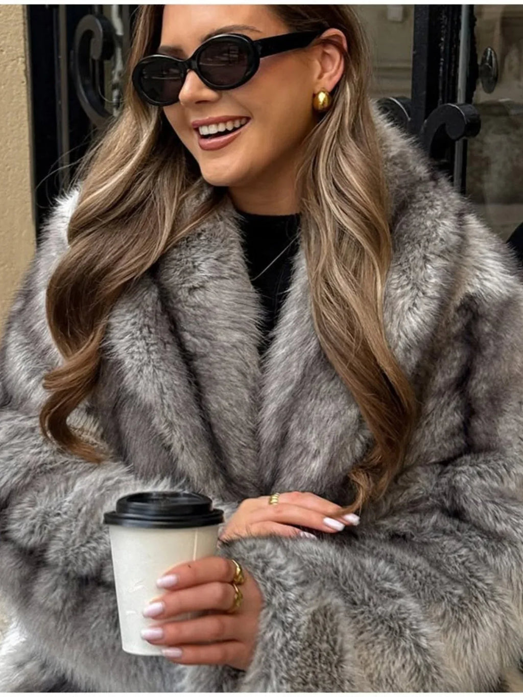 Close-up of a woman's face and upper body wearing a grey faux fur coat, sunglasses, and gold earrings, smiling and holding a coffee cup.
