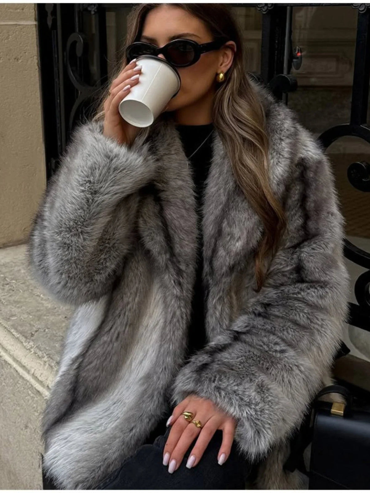 Woman wearing a grey faux fur coat, black top, black jeans, black boots, sunglasses, and gold earrings, holding a coffee cup.