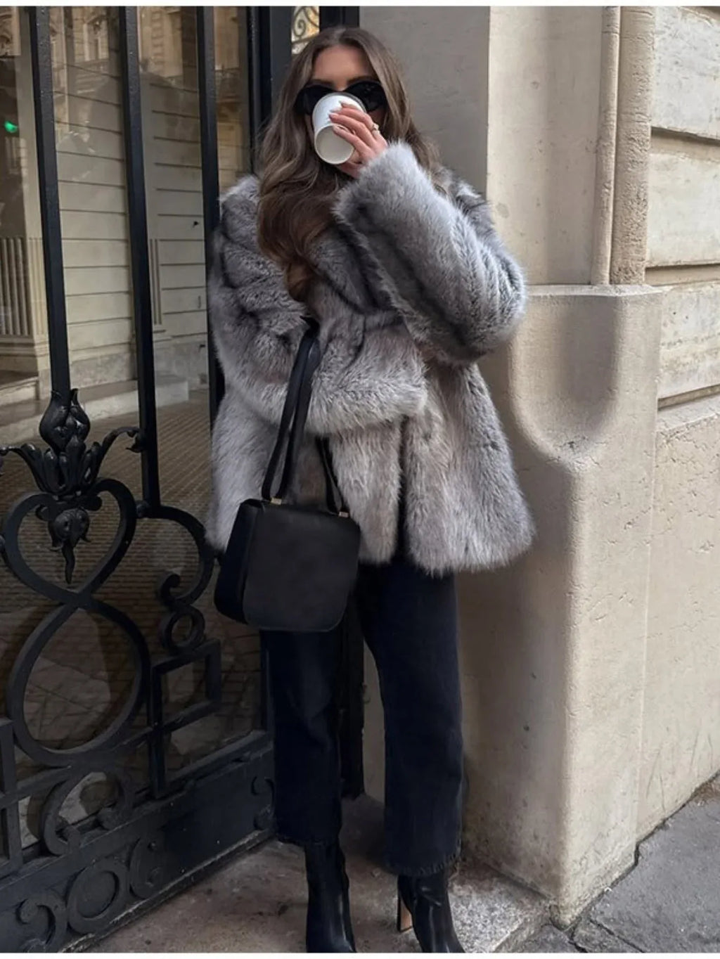 Woman standing outdoors wearing a grey faux fur coat, black top, black jeans, black boots, sunglasses, and gold earrings, holding a coffee cup and a black handbag.