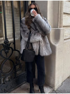 Woman standing outdoors wearing a grey faux fur coat, black top, black jeans, black boots, sunglasses, and gold earrings, holding a coffee cup and a black handbag.