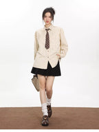 Woman walking towards the camera wearing an oversized beige striped button-down shirt with a brown tie, a black pleated skirt, white socks, and brown loafers, holding a light beige tote bag.