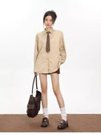 Full body shot of a woman wearing an oversized beige striped button-down shirt with a brown tie, holding a brown tote bag, and wearing white socks and brown loafers.
