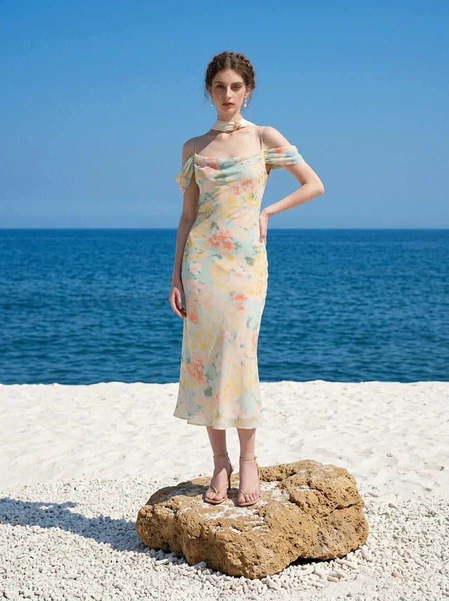 Woman wearing a floral print midi slip dress with off-the-shoulder sleeves and a cowl neckline, standing on a rock on a sandy beach with the ocean in the background.