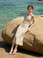 Woman wearing a floral print midi slip dress with off-the-shoulder sleeves and a cowl neckline, sitting on a rock by the sea.