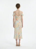 Back view of a woman wearing a floral print midi slip dress with off-the-shoulder sleeves and a cowl neckline, standing against a white background.