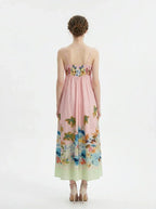 Full back view of a woman wearing a pink midi dress with a tropical print and spaghetti straps, standing against a white background.