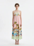 Full front view of a woman wearing a pink midi dress with a tropical print and spaghetti straps, standing against a white background.