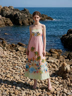 Woman wearing a pink midi dress with a tropical print and spaghetti straps, standing on a rocky beach with the ocean in the background.