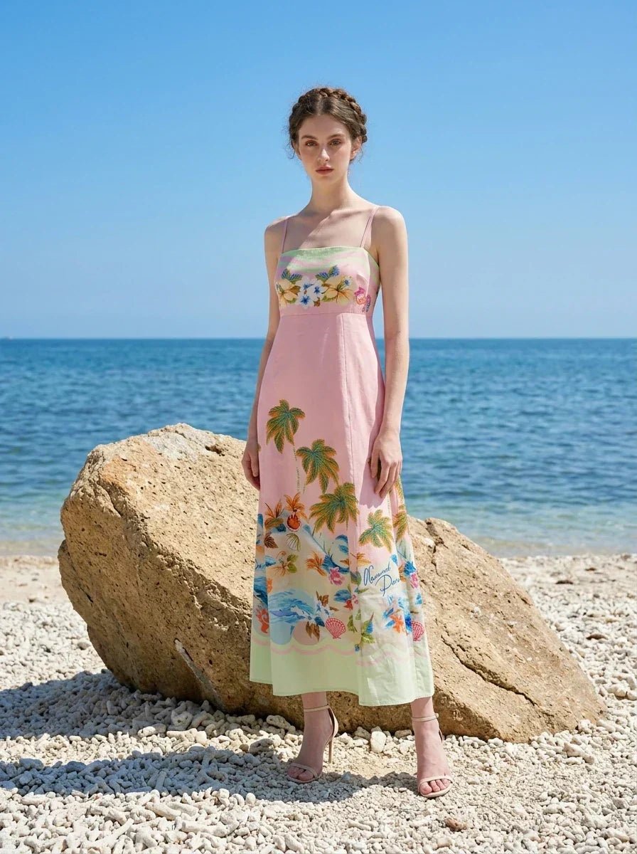 Woman wearing a pink midi dress with a tropical print and spaghetti straps, standing on a beach with large rocks and the ocean in the background.