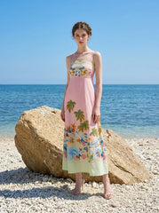 Woman wearing a pink midi dress with a tropical print and spaghetti straps, standing on a beach with large rocks and the ocean in the background.