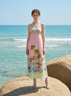 Woman wearing a pink midi dress with a tropical print and spaghetti straps, standing on a rock formation at the beach with the ocean in the background.