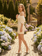 Woman in a white off-the-shoulder satin mini dress standing in a garden path with hydrangeas.
