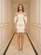 Woman in a white off-the-shoulder satin mini dress standing in a hallway.