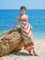 Woman wearing a floral print maxi dress with spaghetti straps, leaning against a large rock on a beach.