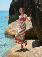 Woman wearing a floral print maxi dress with spaghetti straps, standing on rocks by the sea.