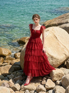 Woman in a red tiered tulle dress with ruffled sleeves and a square neckline, sitting on rocks by the sea