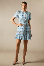 Model wearing a light blue lace sheath dress with short puff sleeves, patch pockets, a belted waist, and a ruffle hem, paired with embellished heels.