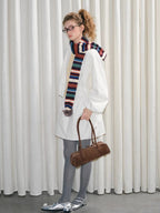 Full body shot of a model wearing a white jacket, gray tights, silver shoes, and holding a brown handbag, with a colorful knit scarf.