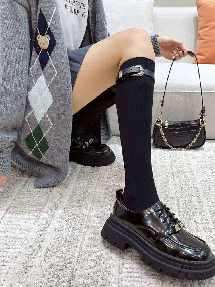 Woman wearing black knee-high socks with adjustable buckle straps, a grey argyle cardigan, and black platform loafers.