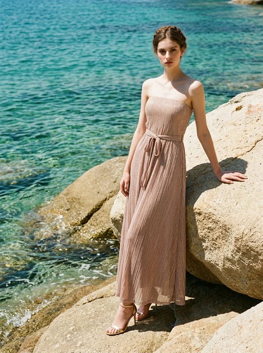 Woman wearing a strapless, metallic knit maxi dress with a self-tie belt, posing on rocks by the sea.