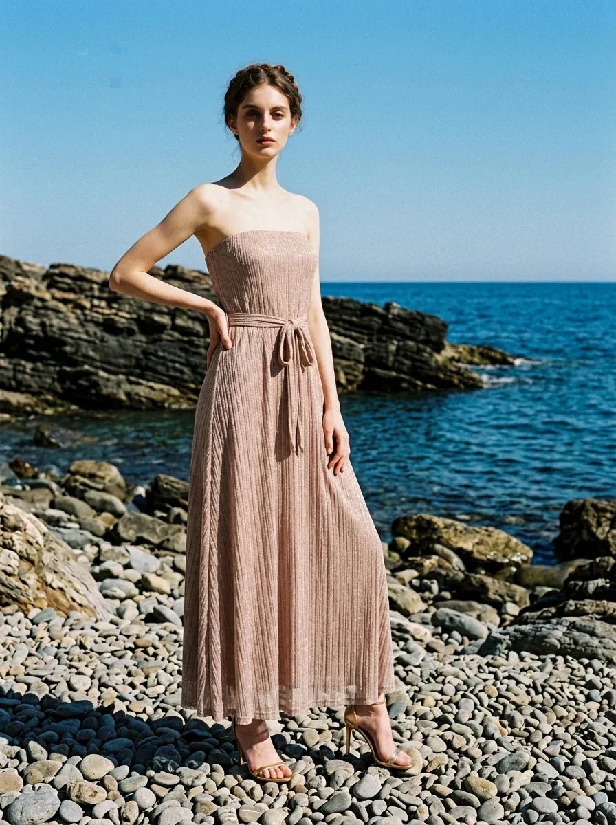 Woman wearing a strapless, metallic knit maxi dress with a self-tie belt, posing on a rocky shoreline with the sea in the background.