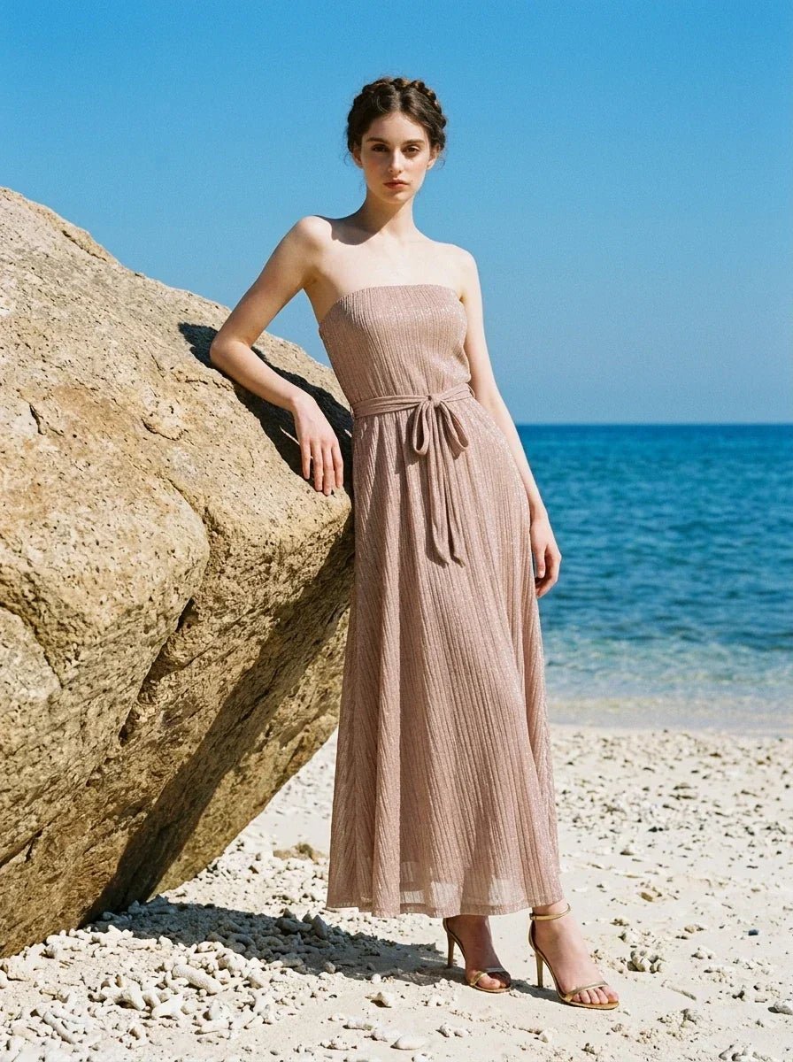 Woman wearing a strapless, metallic knit maxi dress with a self-tie belt, standing on a sandy beach by the sea.