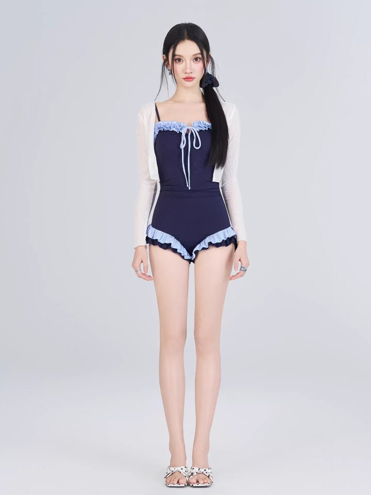 Woman in a navy one-piece swimsuit and sheer white cardigan, standing against a white background.