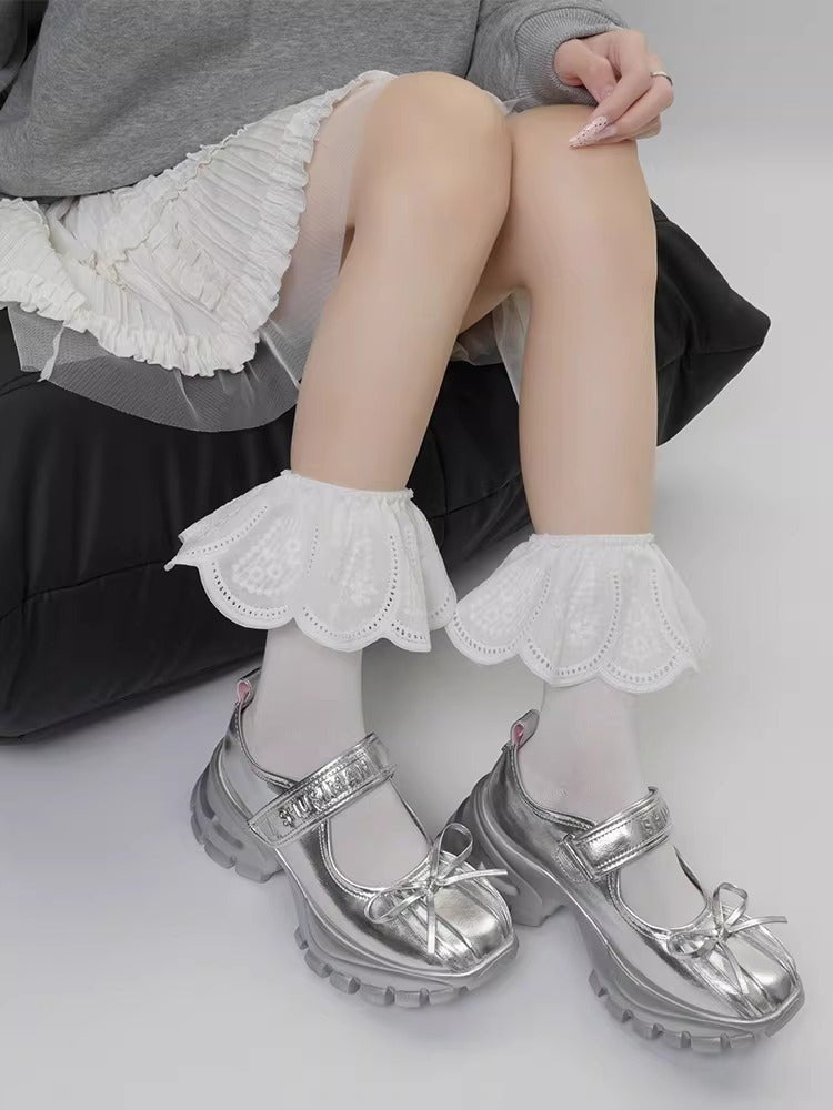 A person wearing white socks with white lace ruffled ankle cuffs and silver platform shoes with bows.