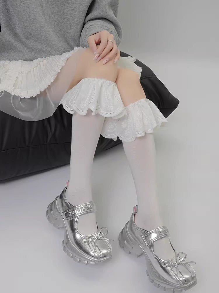 A person wearing white knee-high socks with white lace ruffled cuffs above the knee and silver platform shoes with bows.