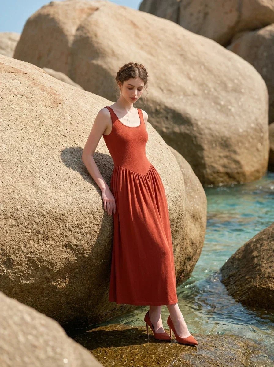woman wearing a rust-colored ribbed knit midi dress with a scoop neckline and sleeveless design, standing near large rocks and the ocean on a beach