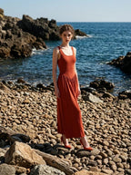woman wearing a rust-colored ribbed knit midi dress with a scoop neckline and sleeveless design, posing on a rocky beach with the ocean in the background