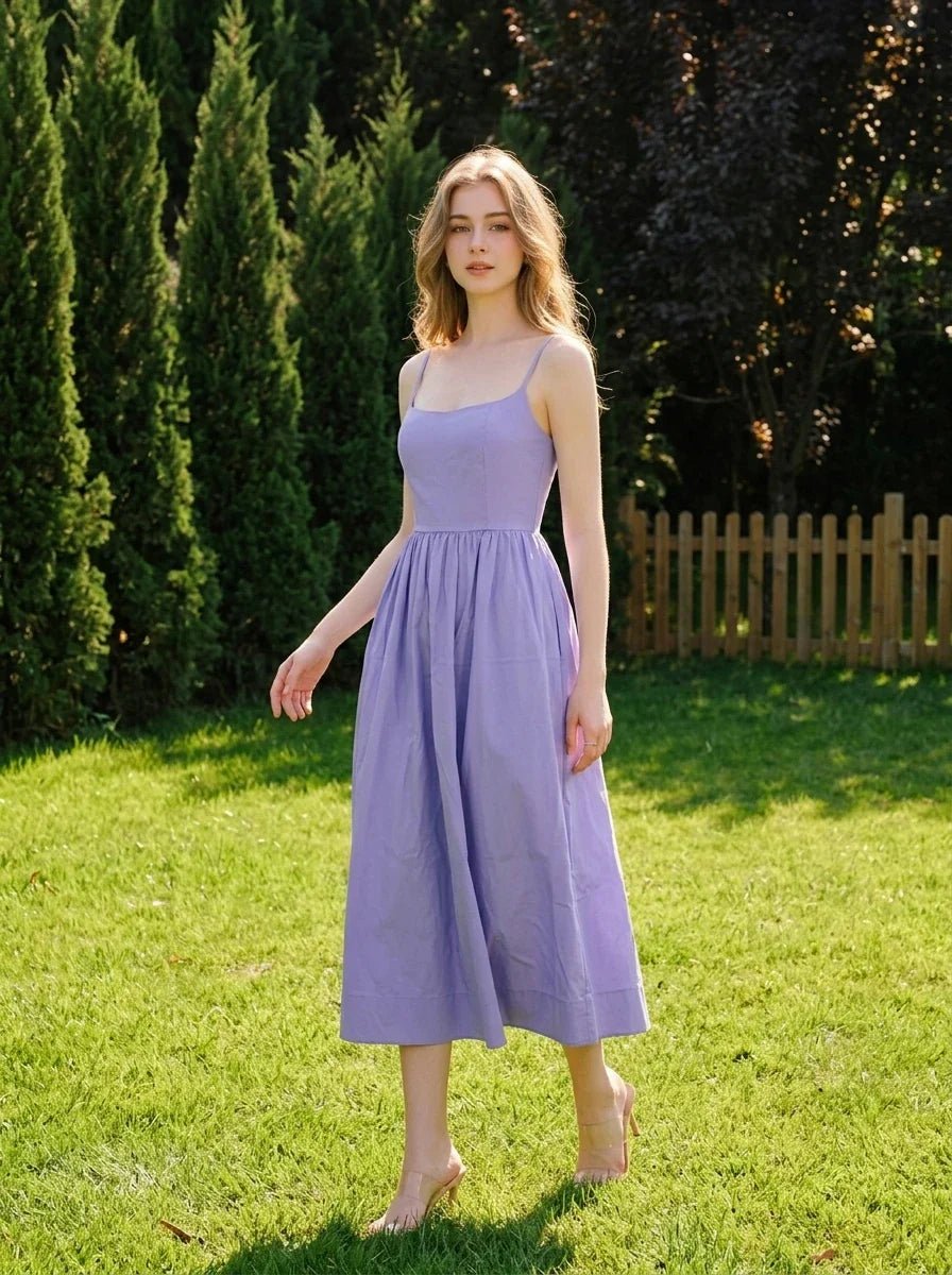 Woman wearing a purple midi dress with spaghetti straps and a square neckline, walking on a lawn in a garden.