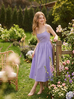 Woman wearing a purple midi dress with spaghetti straps and a square neckline, leaning on a wooden fence in a garden.