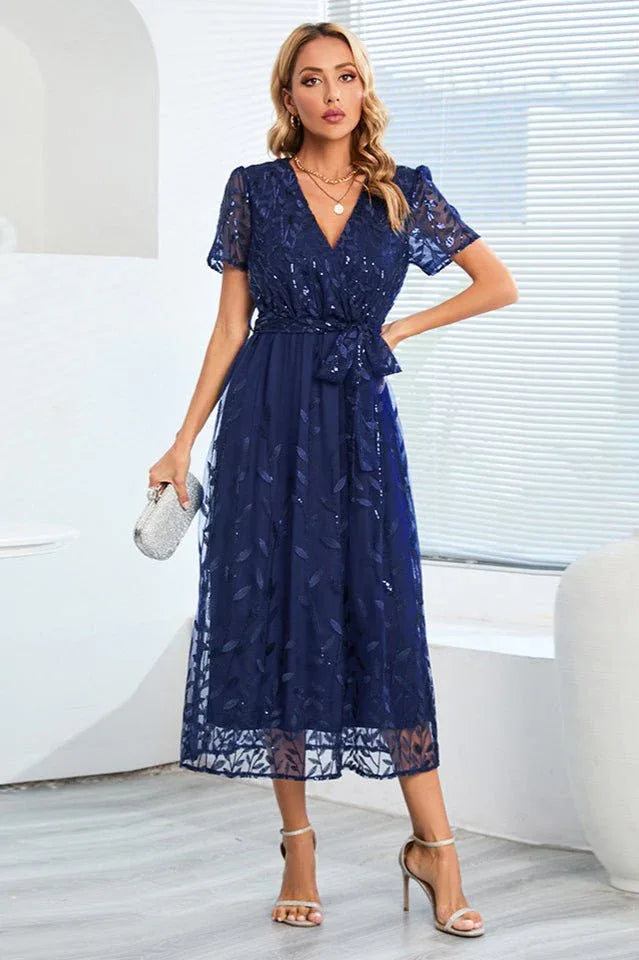 Woman holding a silver clutch and wearing a navy blue sequined midi dress with a leaf pattern overlay. Secondary image