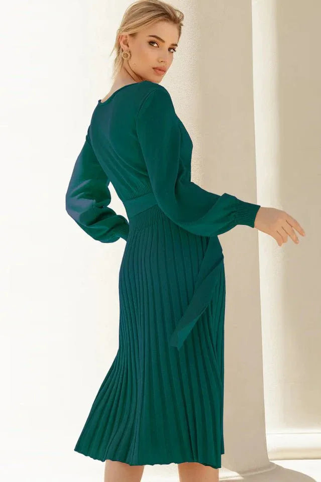Back view of a woman in a long-sleeved green pleated midi dress with a tie waist, turning her head.