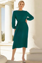 Woman in a long-sleeved green pleated midi dress with a tie waist, standing outdoors with columns in the background.