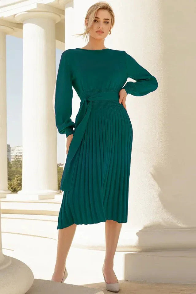 Woman in a long-sleeved green pleated midi dress with a tie waist, standing outdoors with columns in the background.