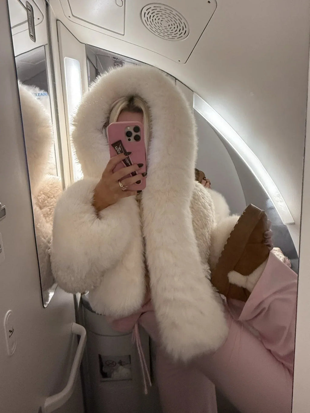 Woman taking a mirror selfie on an airplane, wearing a white faux fur cropped jacket with an oversized hood and pink lounge pants.