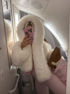 Woman taking a mirror selfie on an airplane, wearing a white faux fur cropped jacket with an oversized hood and pink lounge pants.