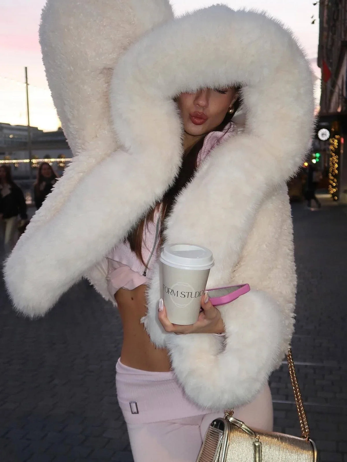 Woman wearing a white faux fur cropped jacket with an oversized hood and pink lounge pants, holding a coffee cup. Main image