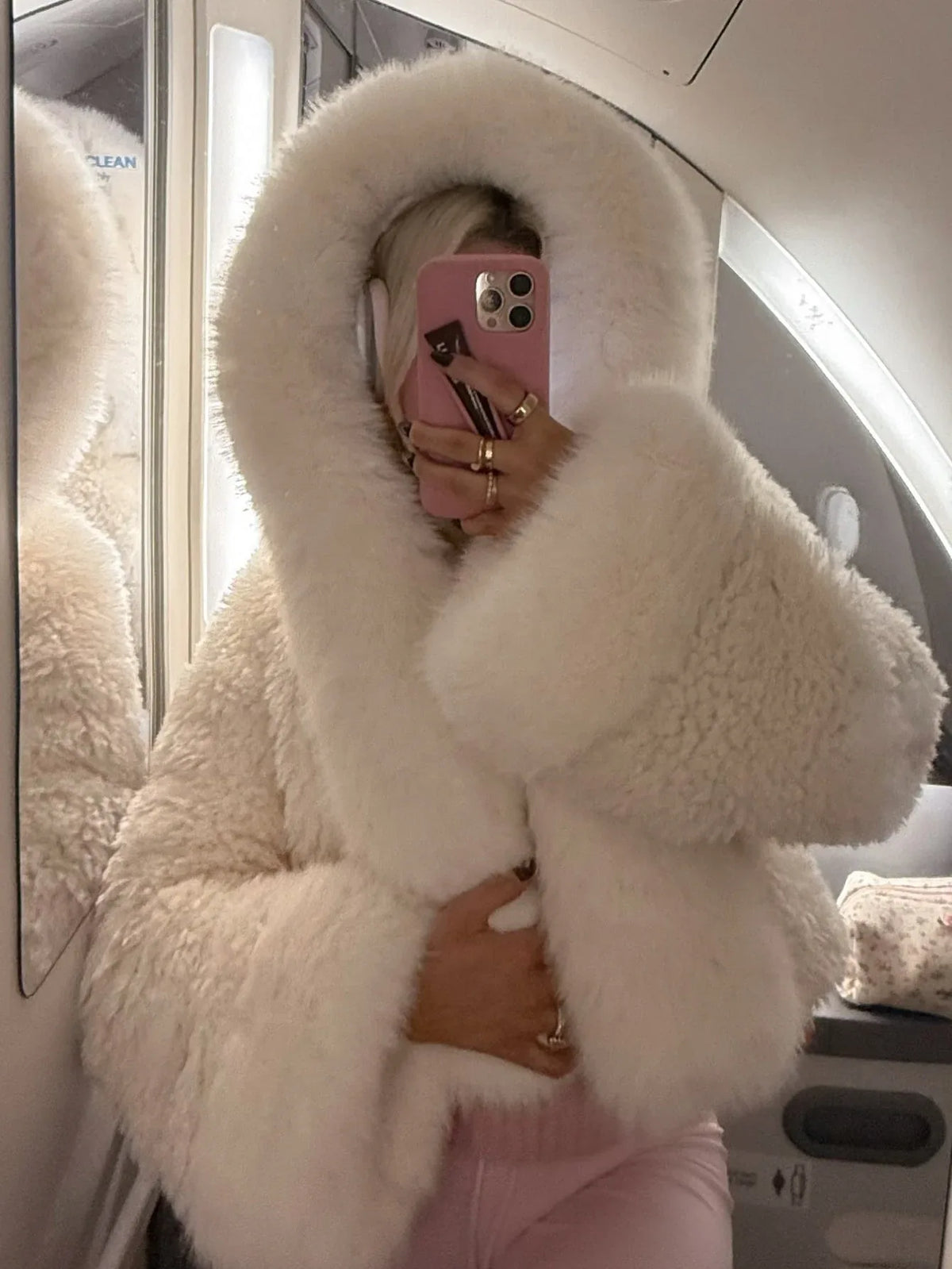 Woman taking a mirror selfie on an airplane, wearing a white faux fur cropped jacket with an oversized hood and pink lounge pants, holding a phone.