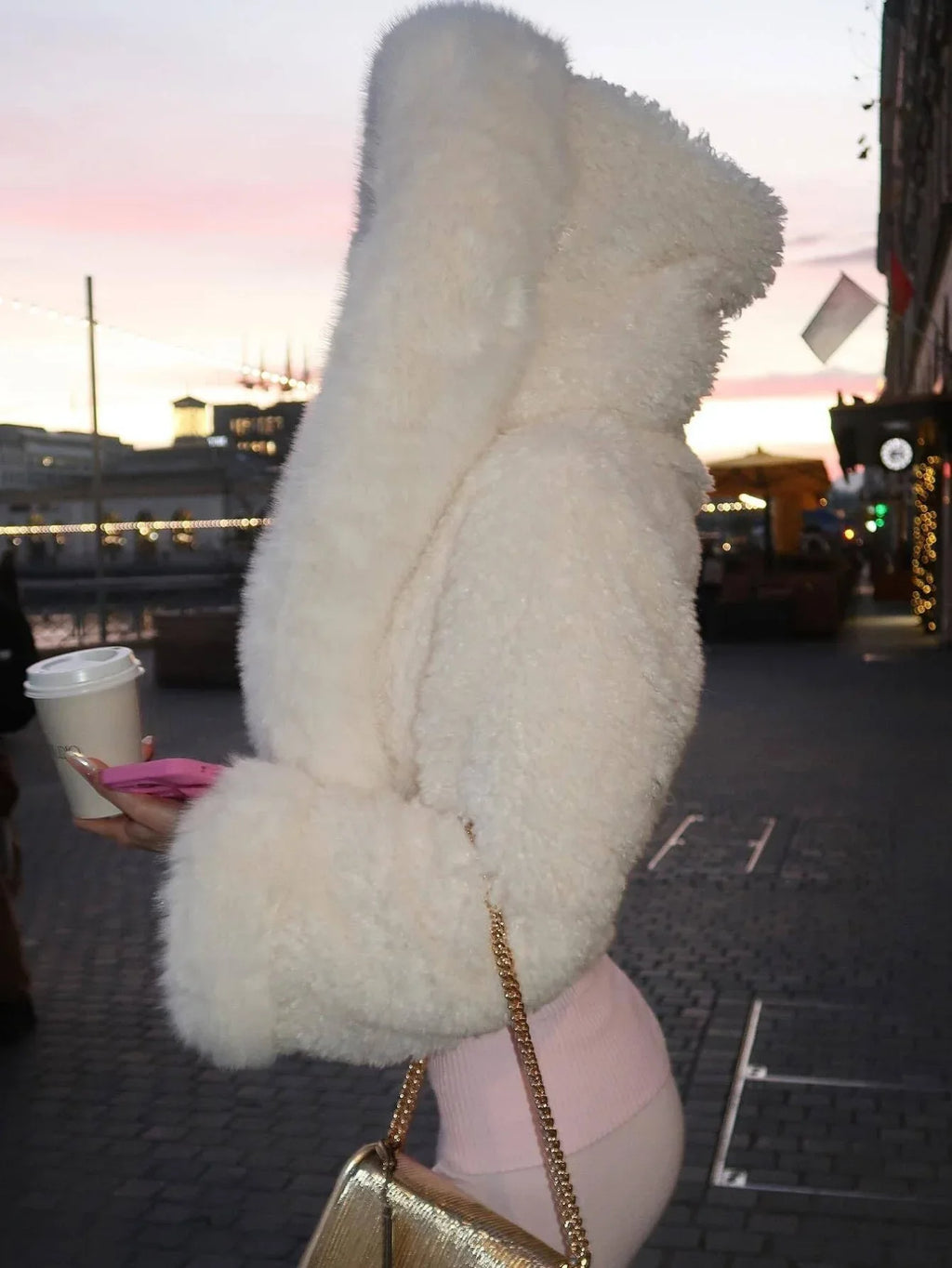 Side view of a woman in a white faux fur cropped jacket with an oversized hood, holding a coffee cup.