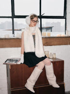 Model sitting on a wooden surface, wearing white fuzzy earmuffs, scarf, gloves, and leg warmers, with a window and candles in the background.