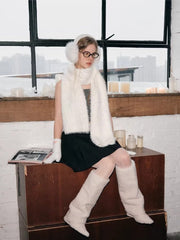 Model sitting on a wooden surface, wearing white fuzzy earmuffs, scarf, gloves, and leg warmers, with a window and candles in the background.