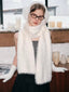 Model wearing a white fuzzy scarf, earmuffs, and gloves with a gray top and black skirt, posing in a studio setting with decorative candles.