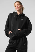 Woman wearing a black cropped hoodie with a kangaroo pocket and embroidered logo.
