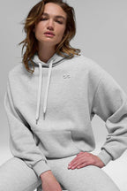 Close-up of a woman wearing a light grey cropped hoodie with a drawstring hood and embroidered logo.