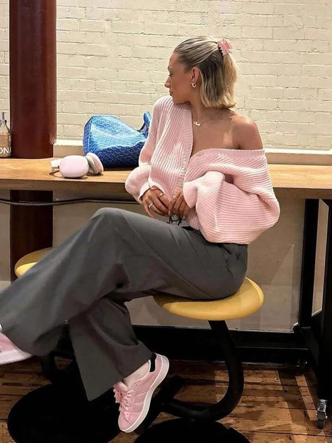 Woman sitting at a table wearing a pink knit cardigan off the shoulder with grey pants.